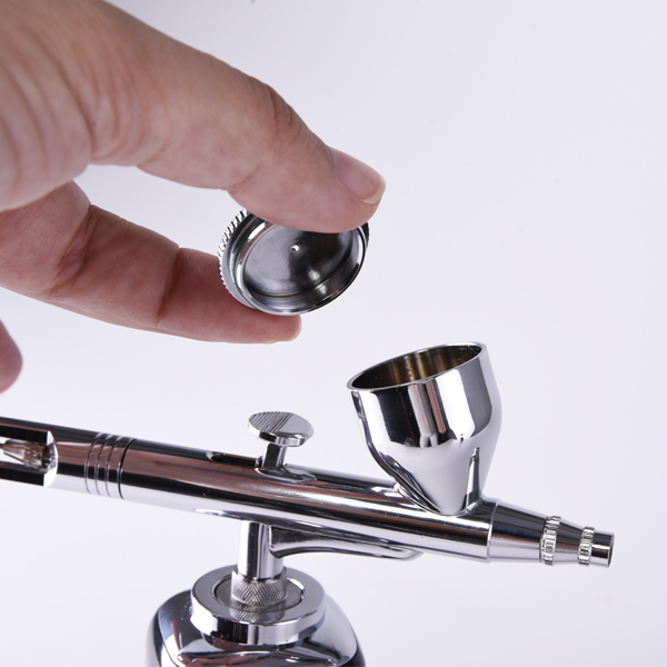 Aerógrafo metálico prateado com copo para tinta e tampa sendo segurada, fundo branco