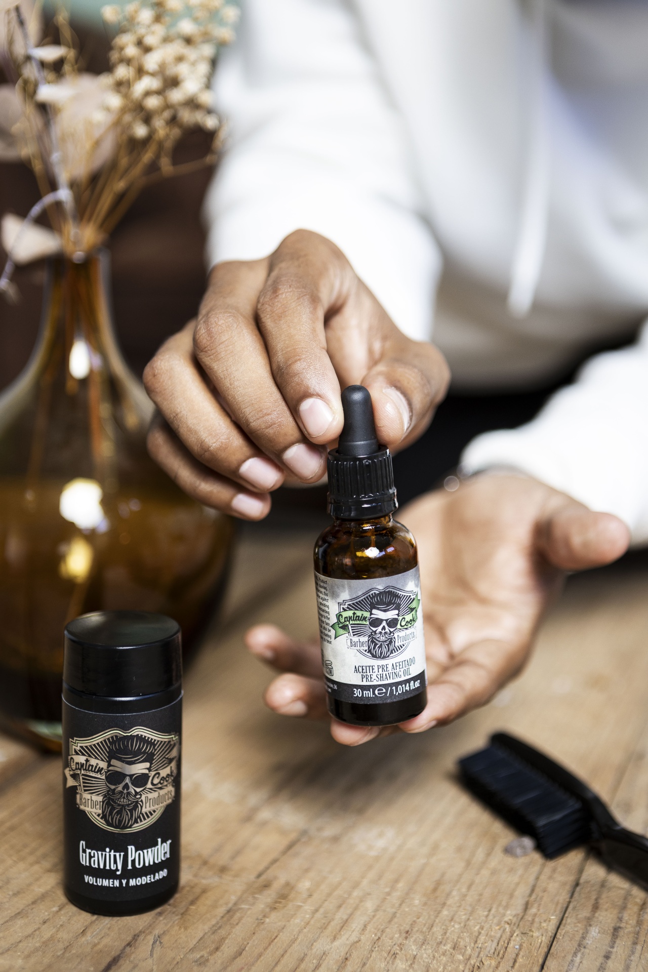 Frascos de óleo para barbear e pó para cabelo sobre mesa de madeira com mão segurando óleo