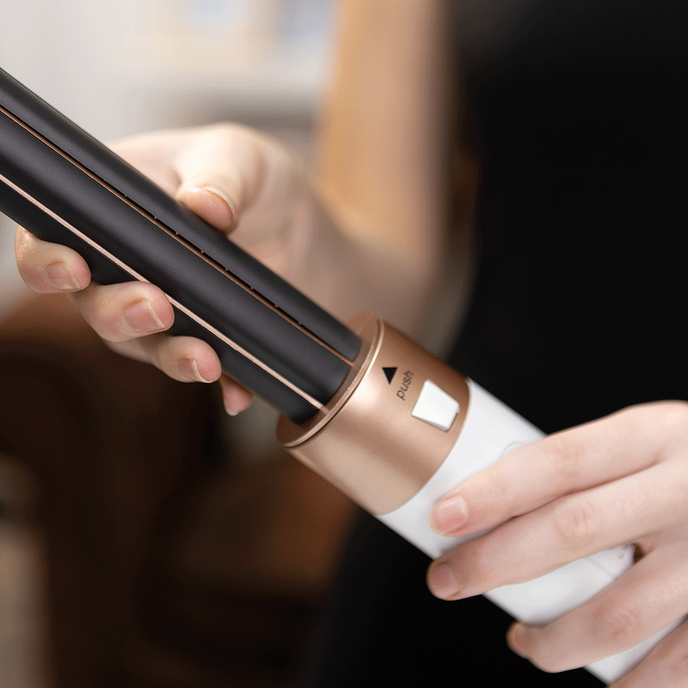 Prancha alisadora de cabelo preta, branca e dourada com botão 'PUSH' sendo segurada por mãos femininas