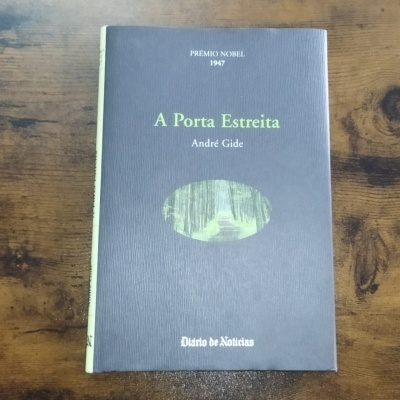 Livro 'A Porta Estreita' de André Gide com capa cinza, texto em verde e branco, e uma pequena imagem oval de uma floresta sobre uma superfície de madeira.