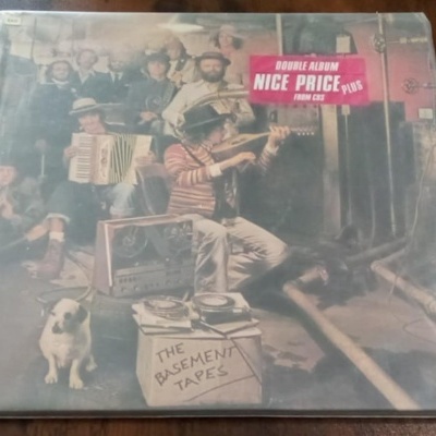 Capa de vinil com várias pessoas, cão branco, aparelhos de som e caixa com texto THE BASEMENT TAPES