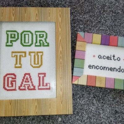 Dois quadros decorativos em ponto cruz com frases em português e molduras de madeira coloridas sobre bancada cinza