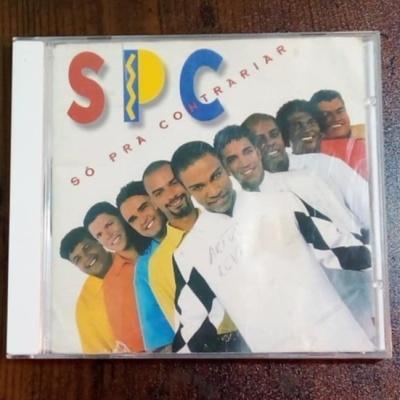 Capa de CD S P C SÓ PRA CONTRARIAR com foto de nove homens em várias cores de roupa, em estojo plástico sobre madeira.
