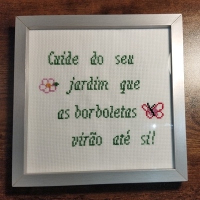Quadro com bordado em ponto cruz com texto em verde e ilustrações de flor e borboleta