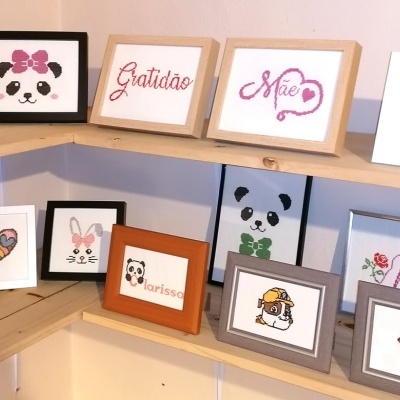 Quadros bordados em ponto cruz com desenhos de pandas, coelhos, corações e texto em português em prateleiras de madeira.