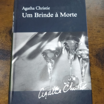 Livro Um Brinde à Morte de Agatha Christie com capa preta e cinza e imagem de copos com líquido