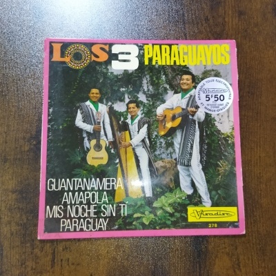 Capa de disco vinil com três músicos vestidos de branco e lenços verdes com instrumentos acústicos em fundo natural