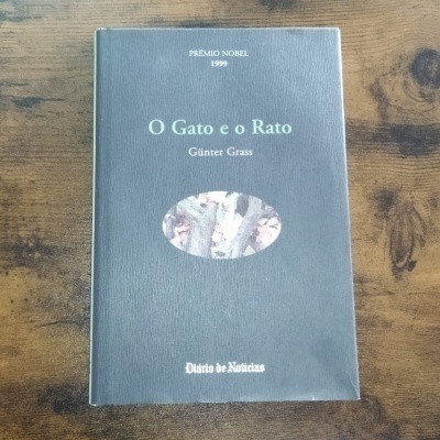 Livro 'O Gato e o Rato' em capa cinza escura com texto branco sobre mesa de madeira