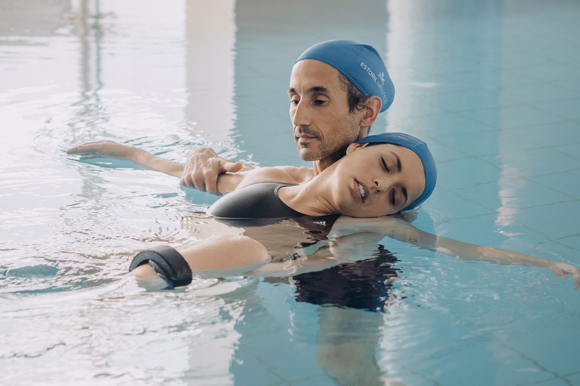 Sessão Watsu (60 min) Dois adultos de touca azul e fatos de banho pretos numa piscina interior, um(a) apoia o outro(a) na água.