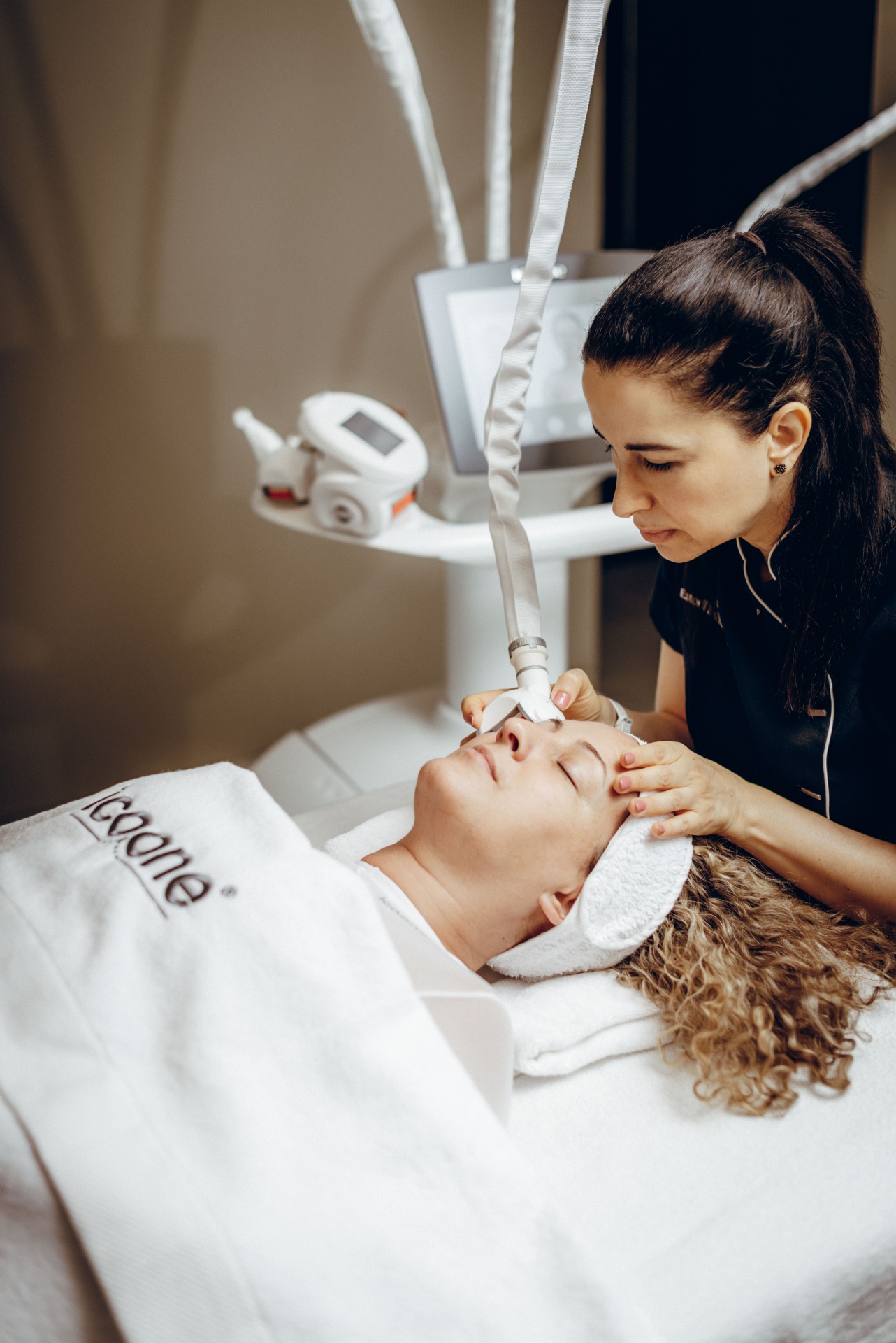 ICOONE® Facial - Tratamento base (30 min) Tratamento facial com aparelho estético em cliente deitada coberta por toalha branca D-Goone