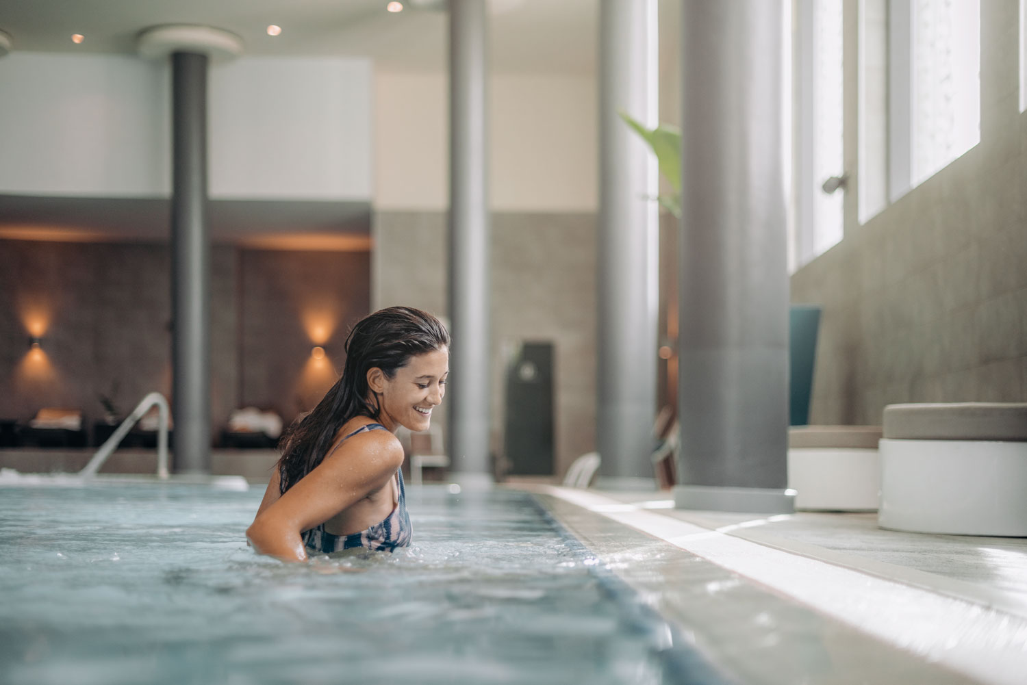 PISCINA DINÂMICA - Circuito HidroBanyan Mulher a entrar em piscina interior com pilares cinzentos