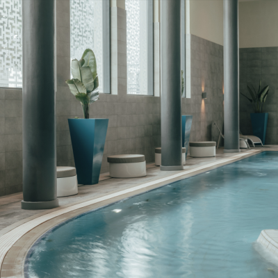 Piscina interior com colunas, plantas em vasos azuis e bancos redondos brancos.
