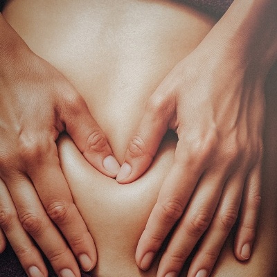 Mãos formando coração sobre pele exposta com pano castanho escuro ao fundo