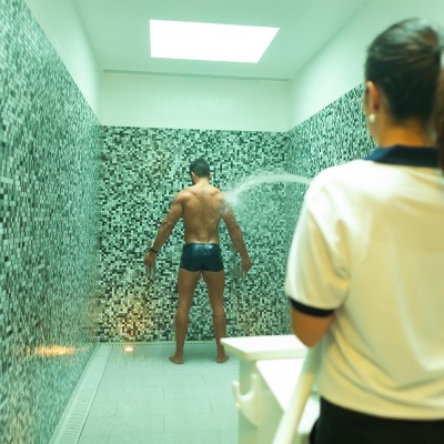 Homem de costas com calção preto numa área revestida de azulejos verdes sendo pulverizado com água por mulher em polo