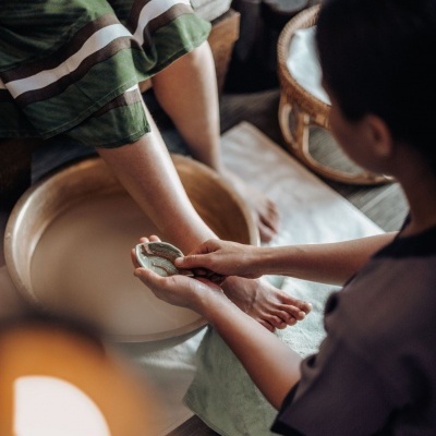Pessoa a fazer pedicure a outra com calças verdes com riscas brancas, pés numa bacia de madeira