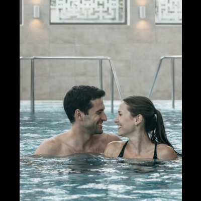 Casal sorrindo dentro de piscina interior com azulejos cinzentos e corrimãos metálicos