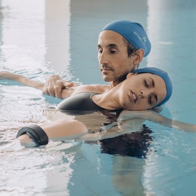 Dois adultos de touca azul e fatos de banho pretos numa piscina interior, um(a) apoia o outro(a) na água.