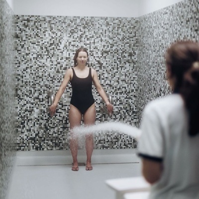 Mulher de fato de banho preto sob chuveiro com pessoas e azulejos quadriculados.