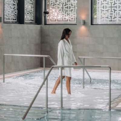 Mulher com roupão branco numa piscina interior com corrimões metálicos e paredes cinzentas