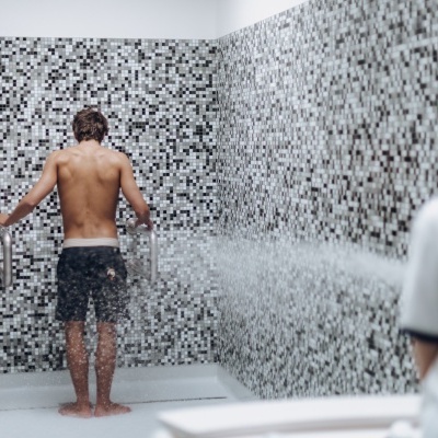 Homem de costas sem camisa num chuveiro com azulejos mosaicos cinza preto e branco segurando corrimões metálicos.