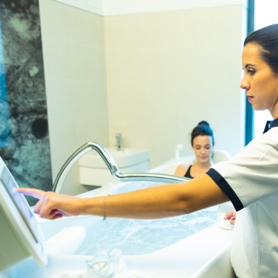 Mulher a usar ecrã tátil num spa com outra mulher na banheira ao fundo