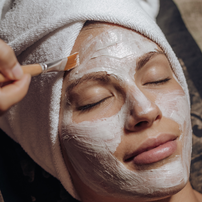 Mulher com máscara facial branca e toalha na cabeça a receber aplicação com pincel