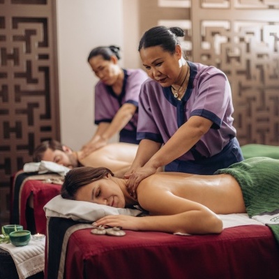 Duas massagistas a fazer massagem em clientes deitadas em macas com lençóis vermelhos e verdes num interior decorado.