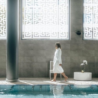 Mulher de bata branca a andar junto a piscina interior com paredes cinzentas e janelas com padrão branco.