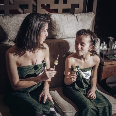 Duas pessoas sentadas em sofá seguram espetadas de frutas com vestes verdes.