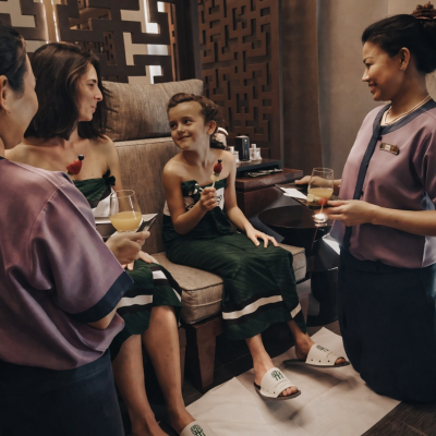 Duas mulheres e uma criança sentadas com roupas de banho verdes e chinelos brancos, servidas por duas funcionárias com uniforme roxo e azul em ambiente acolhedor