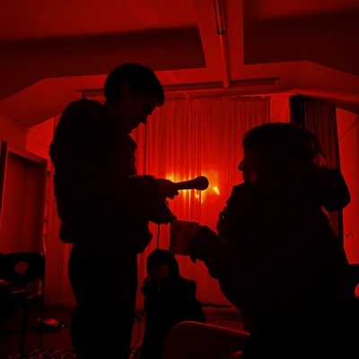Pessoas em silhueta sob luz vermelha intenso com microfone e cortinas ao fundo.