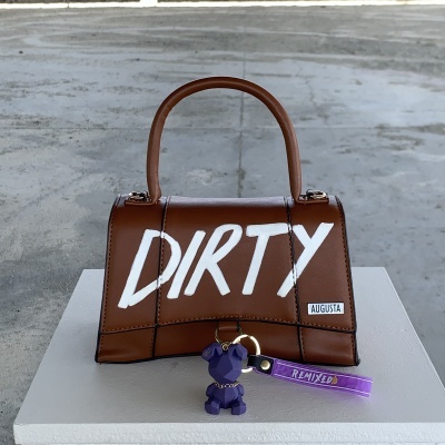 Mala castanha de pele com palavra 'DIRTY' branca, etiqueta 'AUGUSTA' e chaveiro de cão roxo com fita lilás 'REMIXED'