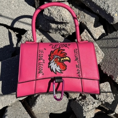 Mala rosa com desenho de galo e texto 'LE COQ' sobre pedras cinzentas