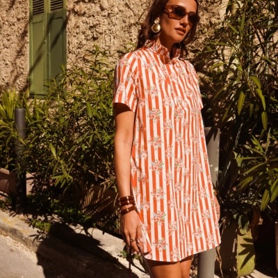 Vestido laranja e branco com flores, menina com acessórios castanhos e óculos escuros numa rua com parede rústica