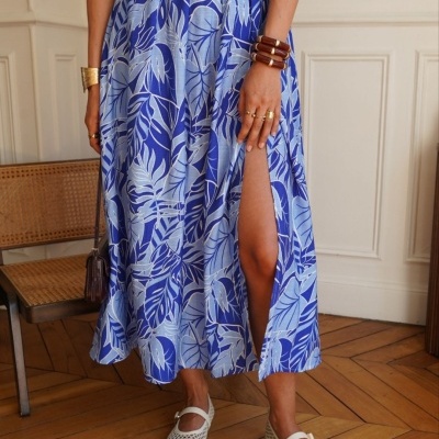 Mulher com saia azul estampada de folhas com racha, camisa branca, pulseiras, anéis e sapatos brancos, em interior com chão de madeira e cadeira de vime