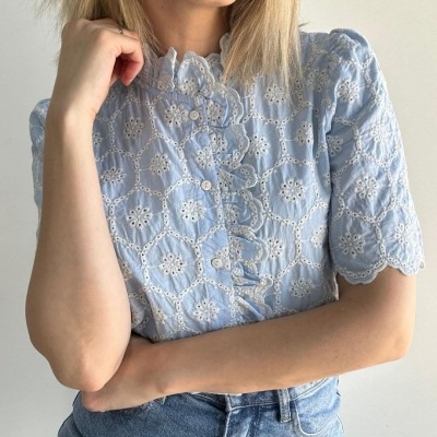 Blusa azul clara bordada com botões e babados, usada com calças de ganga.