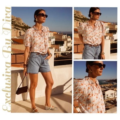 Mulher a usar blusa branca com flores laranja e riscas, calções de ganga, e acessórios dourados, num terraço com vista para edifícios e mar.