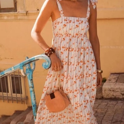 Mulher a usar vestido midi branco com flores laranja e riscas azuis, sandálias brancas, acessórios dourados e castanhos e mala bege