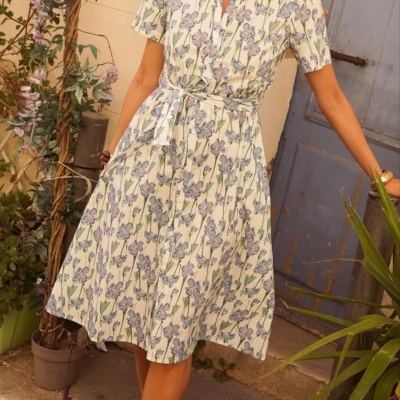 Vestido midi branco com padrão floral azul e verde, com cinto, usado com sandálias azuis, ao ar livre junto a porta azul e plantas.