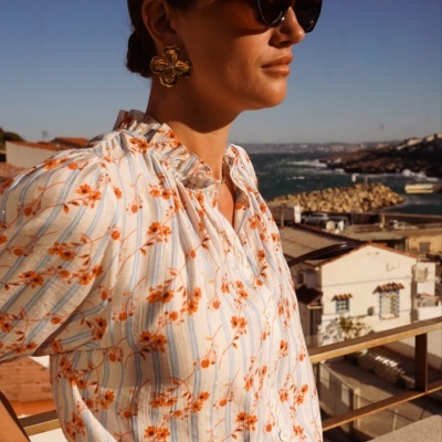 Blusa branca com listras azuis e flores laranja num terraço com vista para o mar.