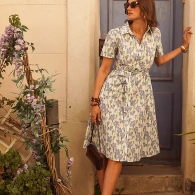 Vestido midi floral branco com cinto, usado por mulher com sandálias azuis, junto a porta azul e plantas em vasos