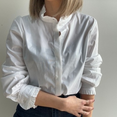 Blusa branca de algodão com folhos na gola e punhos, mangas remangadas e calças de ganga escuras.