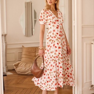 Vestido comprido branco com padrão floral vermelho, mala castanha de pele e sandálias castanhas, mulher num interior com chão em madeira