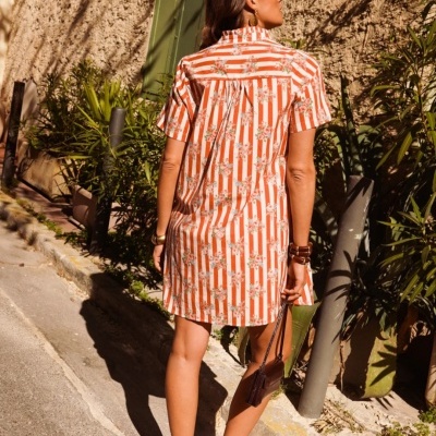 Mulher de costas vestindo vestido laranja às riscas e flores, com acessórios e óculos, em rua com parede texturada e plantas.