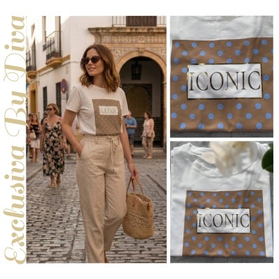 Mulher a usar t-shirt branca com estampa castanha com pontos azuis e texto ICONIC, calças bege e bolsa de palha numa rua de calçada de pedra.