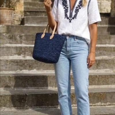 Mulher com blusa branca bordada azul, calças de ganga e mala azul em escadaria.