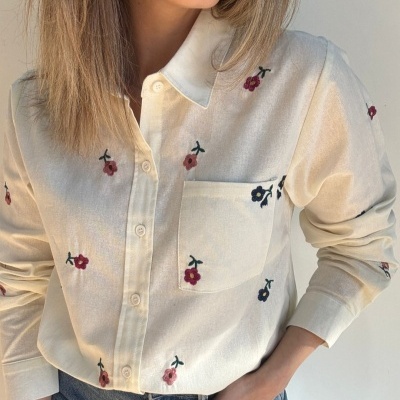 Camisa creme com pequenas flores bordadas, bolso e botões, usada por mulher com jeans azul claro.