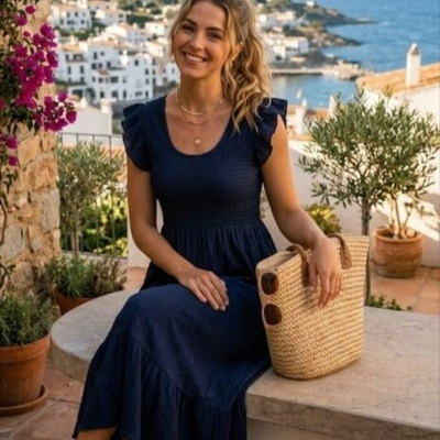 Mulher com vestido azul escuro, sandálias castanhas e mala de palha sentada ao ar livre com vista para casas e mar