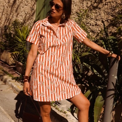 Vestido curto laranja e branco às riscas com flores, mulher com óculos e acessórios, parede texturada e plantas ao fundo