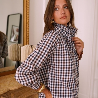 Mulher com camisa de xadrez castanha, preta e branca, tirando a mão do bolso, perto de lareira com moldura dourada e espelho.